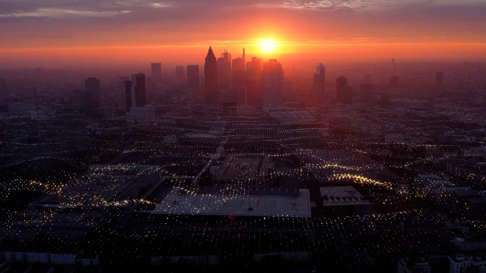 Sunset over a city skyline with a dense network of glowing lights across the foreground.