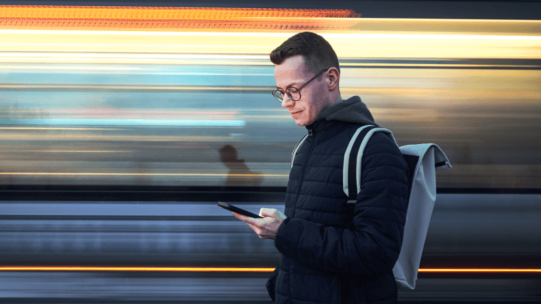 Image   of a person with a backpack looking at a mobile phone while a train appears to zoom by