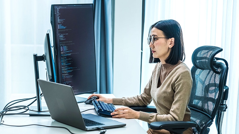 Woman working at a desk with a laptop and a vertical monitor displaying code.